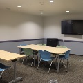 An empty modern collaboration room with light wood tables, blue and green chairs, and a wall-mounted TV.