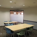 An empty collaboration room with light wood tables, blue and green chairs, and frosted glass windows.