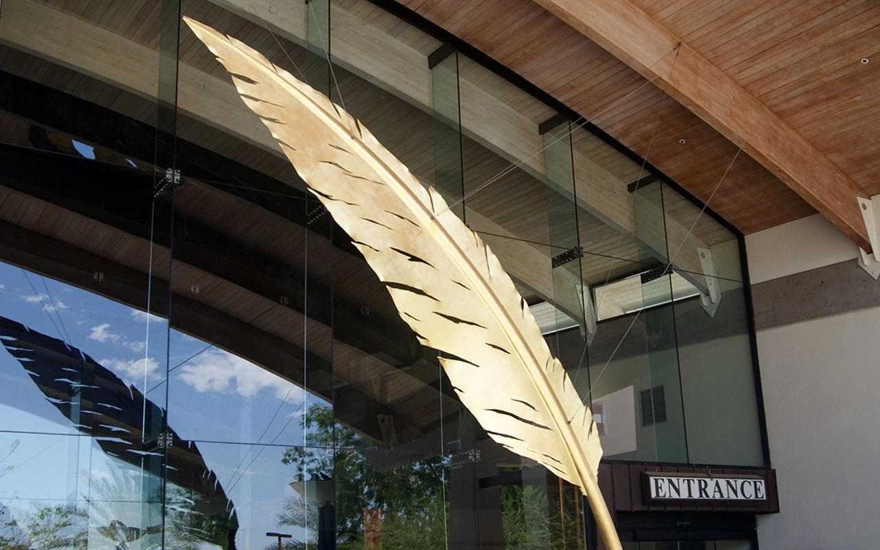 Large golden feather sculpture hanging outside a glass building entrance.